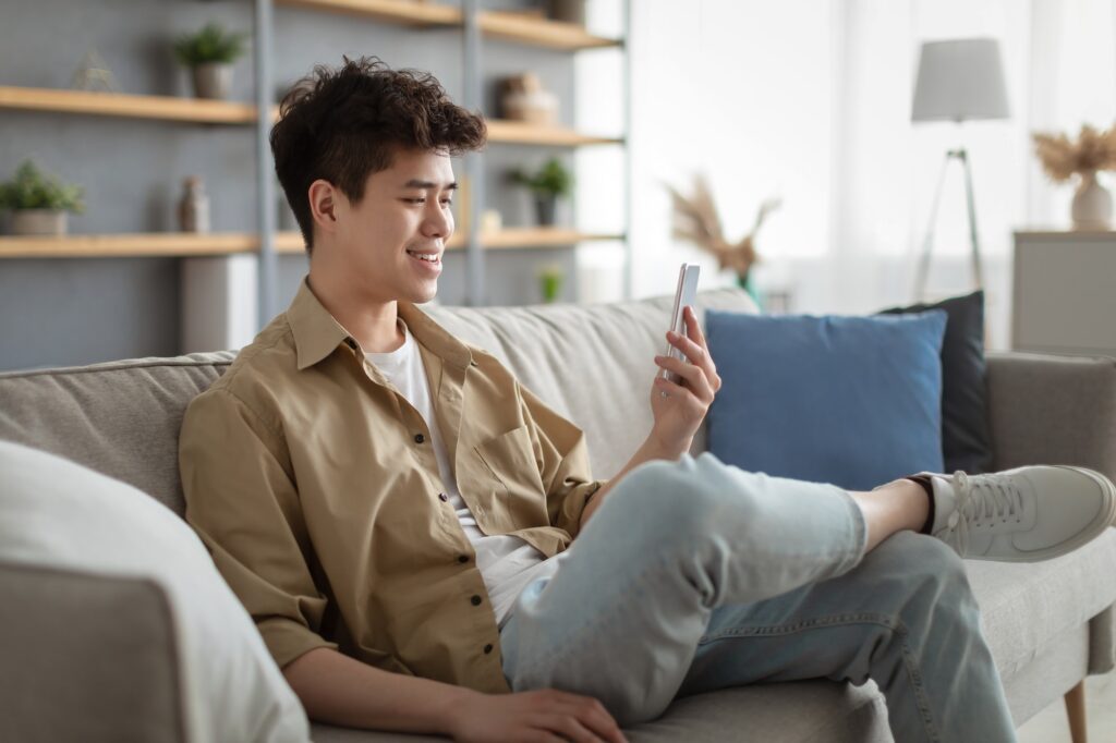 Smiling asian man using smartphone at home sitting on couch